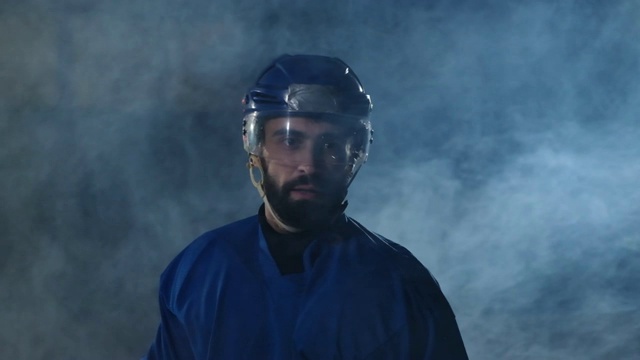 职业冰球运动员拿着一根棒子和一个冰球，在一个黑暗的背景和烟雾中穿着溜冰鞋和头盔在柳达上移动。在冰上场地上用一个年轻人的冰球运球视频素材