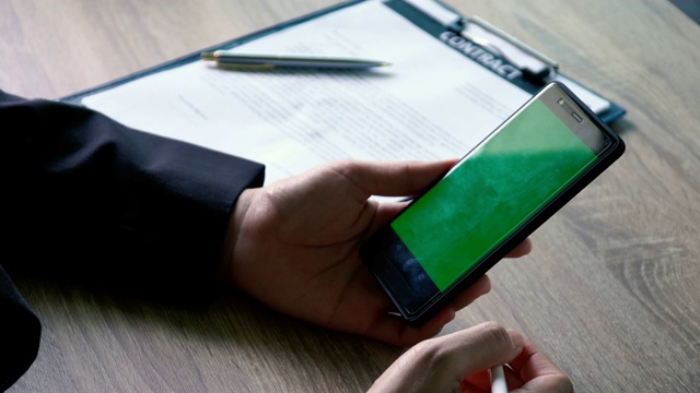 近距离商务女性拿着智能手机滚动和阅读内容的绿色屏幕在办公桌上与合同文件背景视频素材
