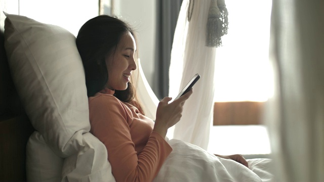 年轻的亚洲女子使用智能手机，早上醒来后躺在床上视频购买