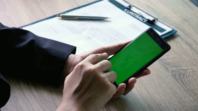 近距离商务女性拿着智能手机滚动和阅读内容的绿色屏幕在办公桌上与合同文件背景视频素材