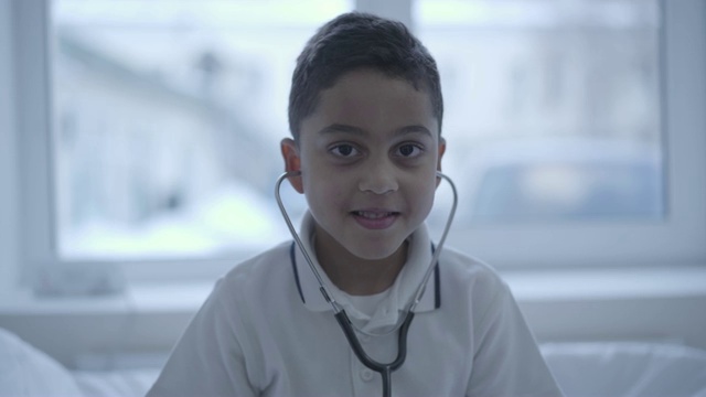 可爱的小男孩的肖像与听诊器在他的耳朵看在相机微笑。视频下载