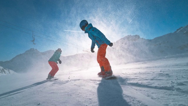 SLO MO TS男性和女性滑雪板骑下一个阳光斜坡和雪颗粒在空中飞行视频下载
