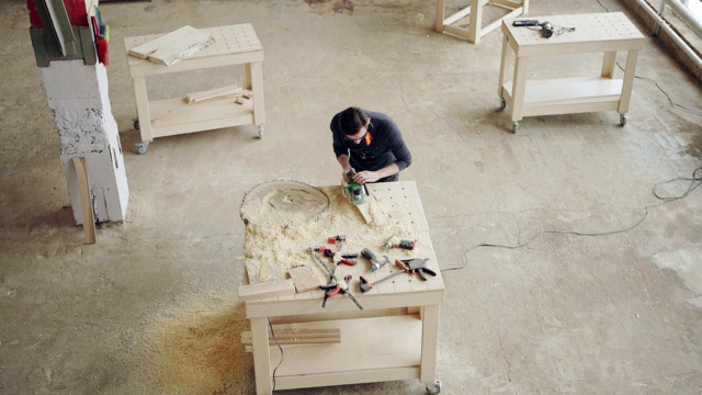 高角度跟踪拍摄工匠在阁楼车间工作的工作台。熟练的木匠用平板砂光机打磨木板，慢动作擦拭锯末视频下载