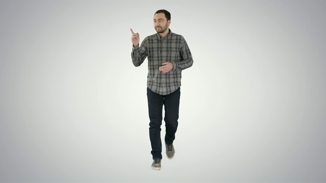 年轻自信的男子在衬衫和牛仔裤走向相机，并指向侧面的梯度背景视频素材