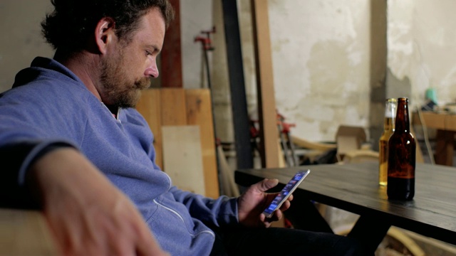 一个留着胡子的男人正坐在桌子前用手机写短信，他面前的桌子上放着两瓶啤酒视频素材