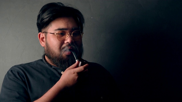 在黑色背景前抽着烟斗的亚洲胡子男子(2)视频素材