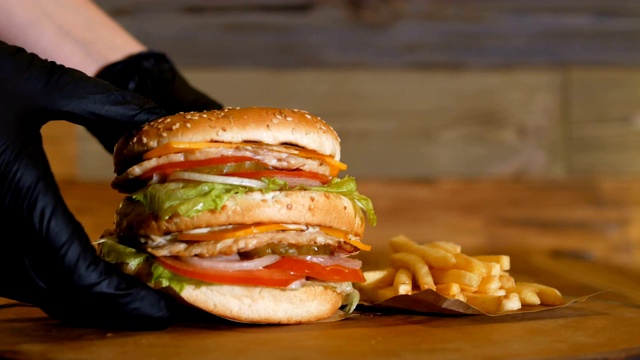 黑手套把一个多汁的汉堡、两片肉片和蔬菜放在薯条旁边的木板上。不健康的快餐。视频下载