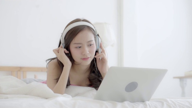 美丽的亚洲女人快乐的耳机听音乐与放松和享受在卧室，女孩好玩的耳机玩笔记本电脑，生活理念，慢动作。视频素材