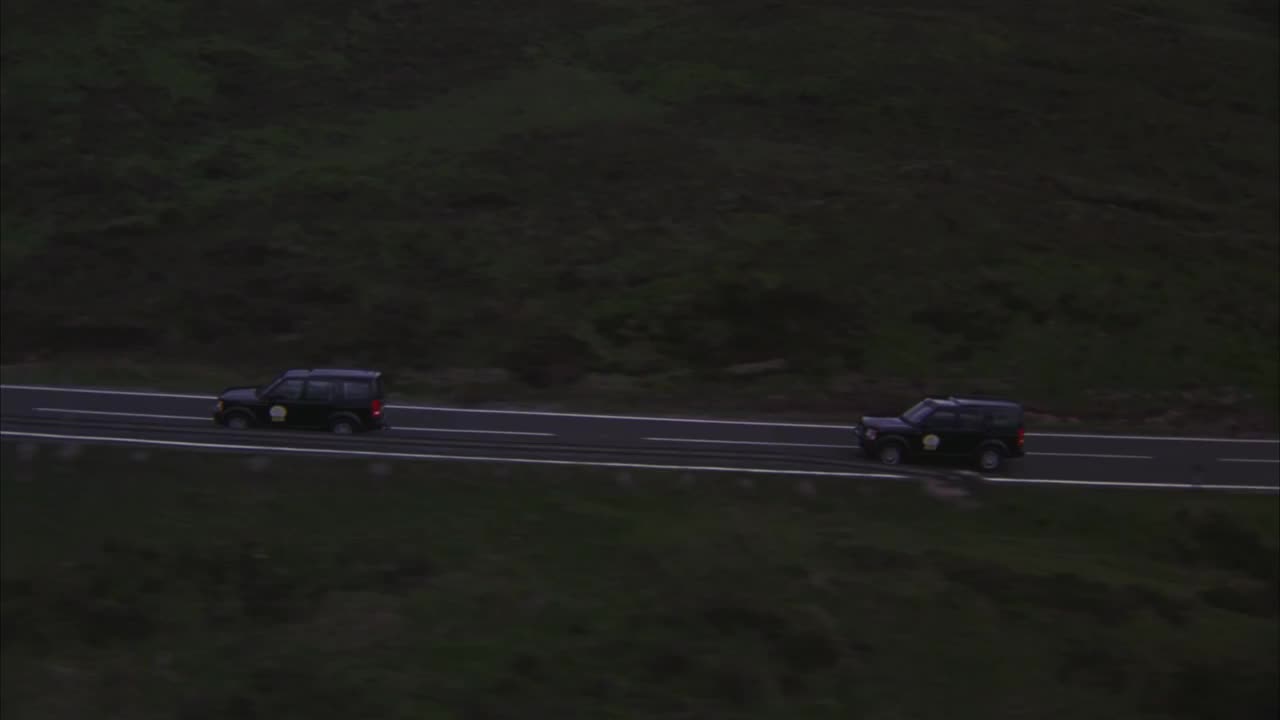 空中跟踪三辆路虎越野车行驶在两条车道的山路上，穿过苏格兰风景中绿色起伏的山丘和山峰。低的乌云。多云或阴天。车队。视频下载