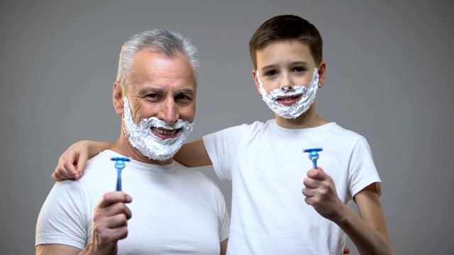 兴高采烈的爷爷和孙子在剃须泡沫中展示剃须刀，男人的仪式视频下载