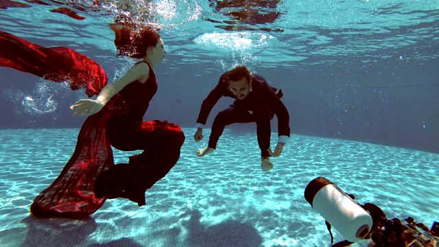 水下摄影师以水面灯光为背景，在泳池底部拍摄穿着婚纱的新娘和新郎。缓慢的运动。视频下载