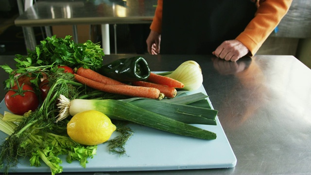 一位年长的白人妇女拿起一个装满蔬菜的切菜板，把它放在一个商业厨房的不锈钢台面上视频素材