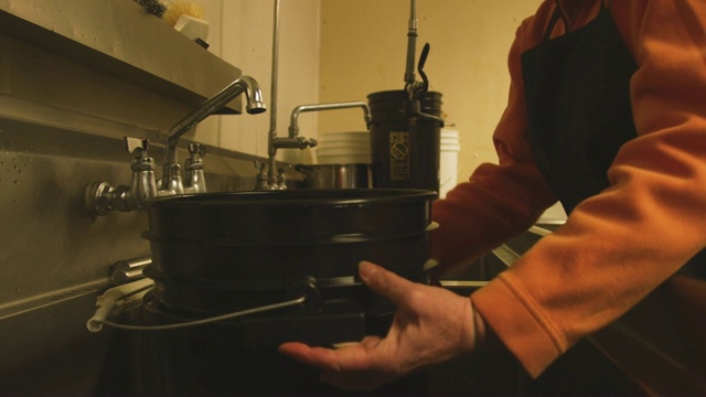 一位年长的白人妇女把一个五加仑的水桶放进一个商业水槽，然后在一个商业厨房/制造设施里装满水视频素材