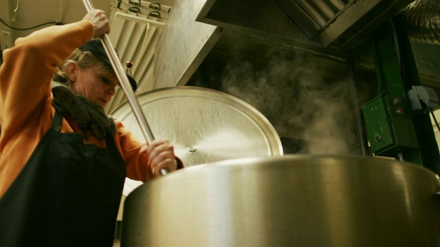 一名60多岁的白人妇女在一个商业厨房/制造设施中，用长桨在一个大型金属储存罐中搅拌液体食物视频素材