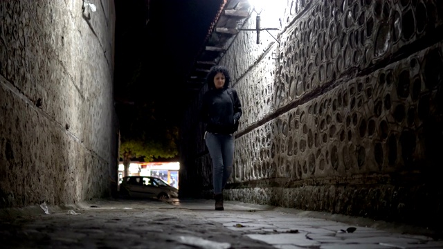 女人在牛仔裤和靴子走在黑暗的城市小巷在晚上，电影低角度拍摄视频素材