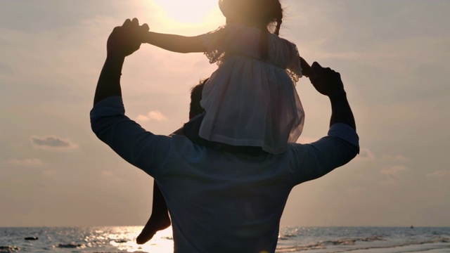 亚洲女婴的剪影骑在父亲的脖子上。在一个夏天的日落时分，爸爸和小女孩一起在户外玩耍。一家人在海滩上看日落。假日旅行的概念。假期——iStock视频素材