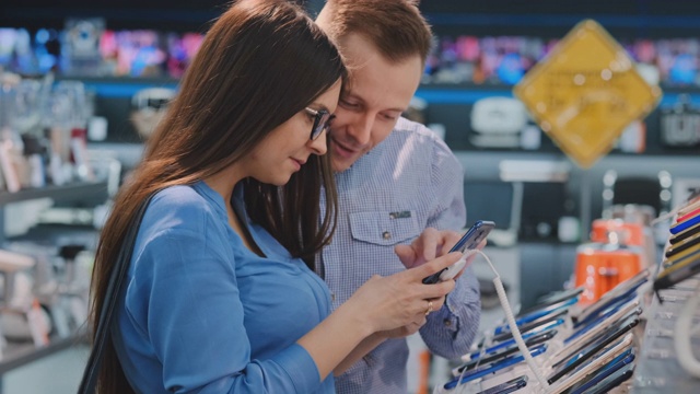 年轻美丽的夫妇。男人和女人手里拿着一部智能手机，站在电器商店展示智能手机的橱窗旁边，挑选最好的智能手机。给他妻子的礼物视频素材