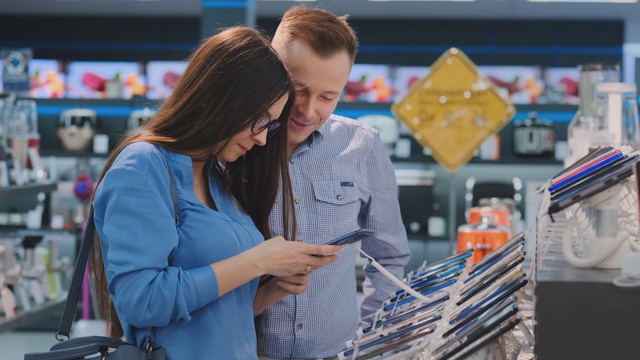 一对年轻漂亮的已婚夫妇，一个穿着衬衫的男人和一个戴眼镜的女人拿着智能手机站在电子产品商店的智能手机展示台上，检查着屏幕的触摸屏，检查着设备的形状和设计视频素材