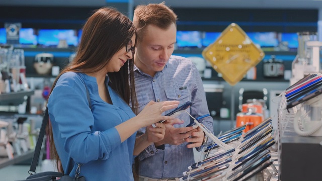 在一家电子产品商店里，一男一女手里拿着两部手机站在展示台上，从两款最好的手机中挑选。两个设备的比较。产品选择多视频素材