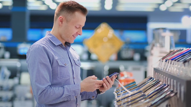 一名拿着智能手机站在商店橱窗前的男子在一家便携式电子产品商店选购一款新手机。购买最新版本的小工具视频素材