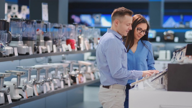一对年轻漂亮的已婚夫妇，一个穿着衬衫的男人和一个戴眼镜的女人拿着智能手机站在电子产品商店的智能手机展示台上，检查着屏幕的触摸屏，检查着设备的形状和设计视频素材