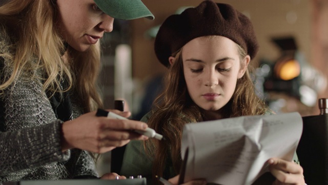 女制片人和一个年轻的女孩坐在导演的椅子上看拍摄脚本，并在电影片场做笔记。视频素材