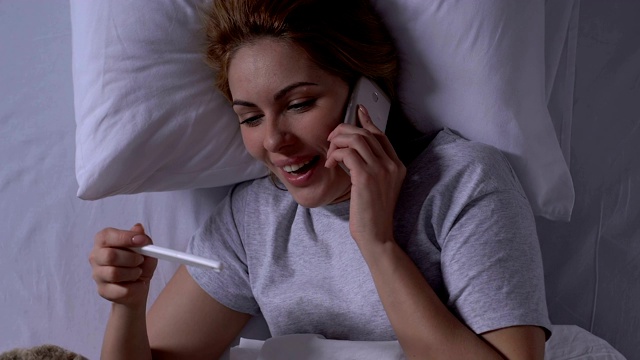 微笑的女人在床上抱着验孕棒和打电话，结果是阳性视频素材