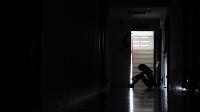 一个悲伤的年轻女孩的剪影坐在黑暗中靠在旧公寓的墙，家庭暴力，家庭问题，压力，暴力，抑郁和自杀的概念。视频素材