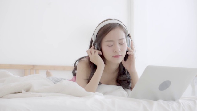 美丽的亚洲女人快乐的耳机听音乐与放松和享受在卧室，女孩有趣的耳机玩笔记本电脑，生活方式的概念，慢动作。视频素材