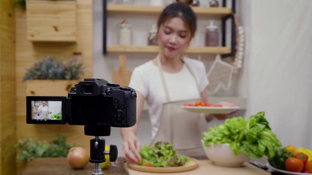 年轻的亚洲女性美食博主说话，同时录制视频准备绿色沙拉在厨房，教程视频到社交网络通过互联网在家概念，选择性焦点视频素材