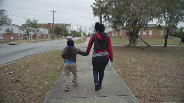 镜头跟随年轻的非洲裔美国母亲和儿子手牵着手走在社区的人行道上。视频下载