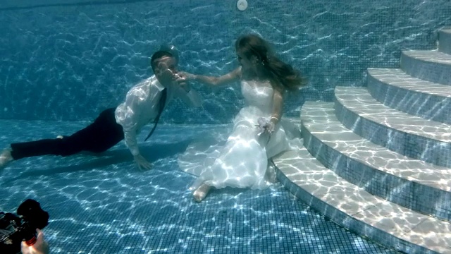 新娘和新郎在水下拍照。身穿白色婚纱的新娘坐在池底，新郎亲吻她的手。在水下婚礼。操作相机。4 k。视频下载