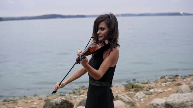 美丽的女孩在黑色的裙子拉小提琴在海上的背景。艺术概念4k视频下载