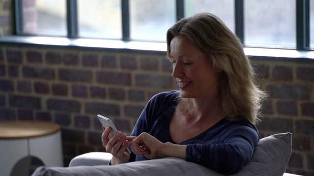 白种人美丽的女人在家里放松在沙发上聊天用智能手机微笑视频素材