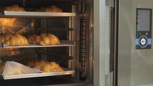 一位专业的面包师打开专业的加热烤箱，拿出盛有新鲜出炉的热羊角面包的托盘。牛角面包在烘焙屋的制造。糕点用品、面包店、糕点店。视频下载