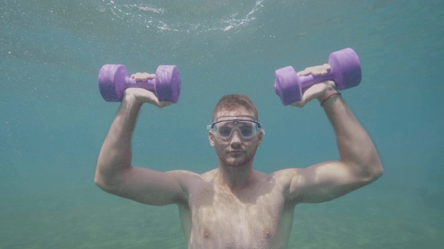 年轻的运动运动员戴着面具在水下用哑铃练习肩膀，正面观看。视频下载