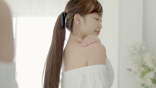 美丽的年轻亚洲女性照镜子和应用乳液防晒霜的肩膀，美丽的女孩完美的皮肤护理和健康与霜治疗和保湿，健康和保健的概念。视频素材