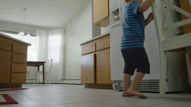 一个三岁的白人男孩走进厨房，把椅子滑到一个柜子上，然后爬到椅子上从柜子里拿东西视频下载