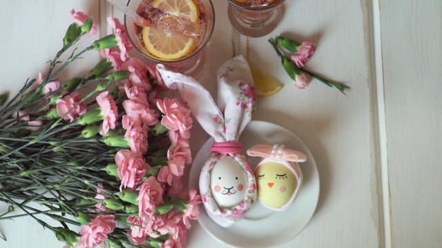 女手放有复活节彩蛋的盘子，装饰着复活节兔子和复活节小鸡，背景是粉红色的康乃馨和柠檬水视频素材