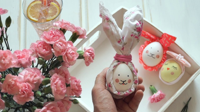 手持复活节彩蛋装饰复活节兔子耳朵和油漆口吻，以装饰彩蛋，粉红色康乃馨和柠檬水为背景视频素材