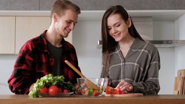 一对穿着格子衬衫的年轻漂亮夫妇正在厨房里做饭。一个女人和一个男人切蔬菜，在一个透明的玻璃盘子里用胡椒、番茄和萝卜做沙拉。视频素材