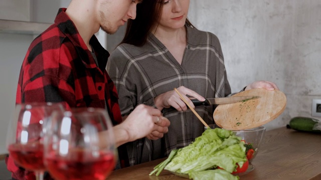 一对穿着格子衬衫的年轻漂亮夫妇正在厨房里做饭。一个女人和一个男人切蔬菜，在一个透明的玻璃盘子里用胡椒、番茄和萝卜做沙拉。视频素材