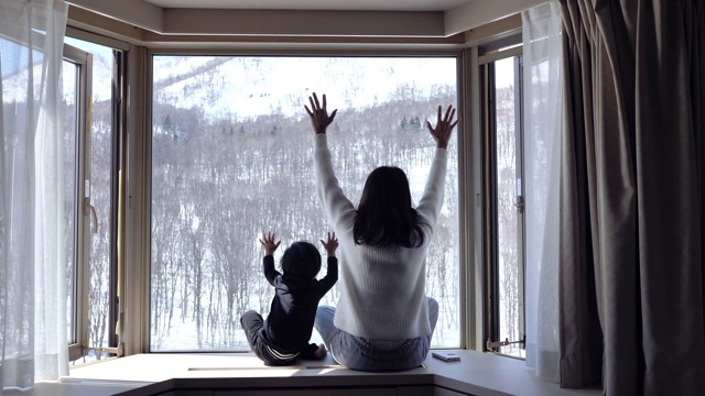 一个小孩和他的母亲坐在窗边，看着森林里的雪花飘落。视频购买