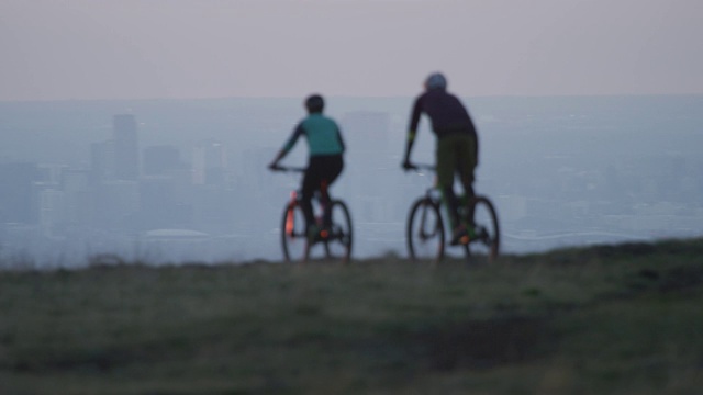 两名骑手(一名女性和一名男性)山地自行车在洛基山脉以西的丹佛，科罗拉多州与城市在日出视频素材