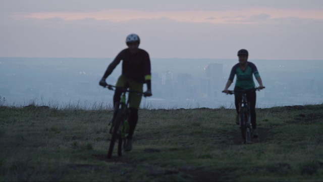 两名骑手(一名女性和一名男性)山地自行车在洛基山脉以西的丹佛，科罗拉多州与城市在日出视频素材
