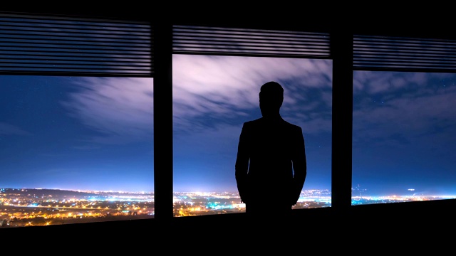 这个男人站在窗户旁边，背景是城市的灯光。时间流逝视频素材