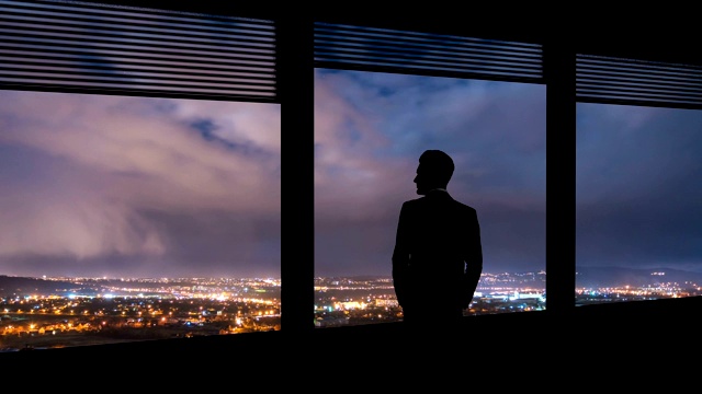 在夜色中的城市背景下，那个商人站在窗边。时间流逝视频素材