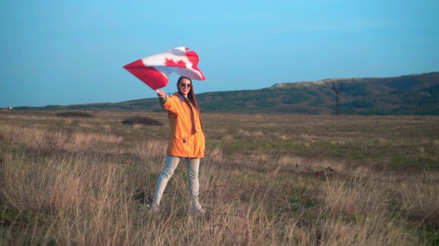 一个穿着黄色夹克，戴着眼镜的女孩手里拿着加拿大国旗。加拿大的国旗正在风中飘扬视频下载