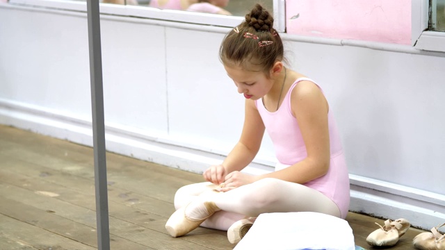 十几岁的小女孩穿着粉红色的紧身衣，准备在芭蕾舞学校的古典芭蕾舞蹈课。她穿着尖头鞋，坐在旧木地板上视频素材