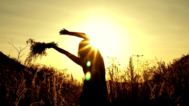 一个年轻的女孩站在大自然在日落，她的手拿着鲜花，把它们举过她的头，从一边到另一边在明亮的夕阳光线。轮廓。的概念。关闭了。4 k。视频下载